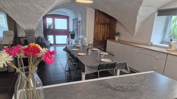 Un comedor con una mesa preparada para cuatro personas, un jarrón con flores y una cocina al fondo.
