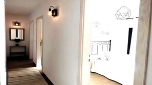 Een hal met een spiegel, een tafeltje met een plant en een deur naar een slaapkamer met een bed en een geometrische wandversiering.
