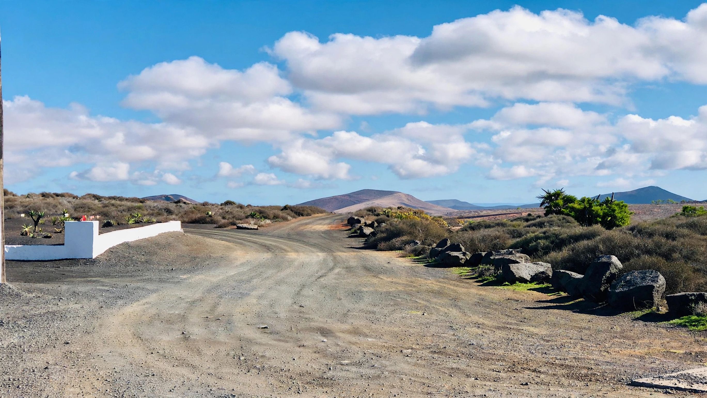 Ein unbefestigter Weg zieht sich durch eine trockene Landschaft mit vereinzelter Vegetation, im Hintergrund ein Berg.
