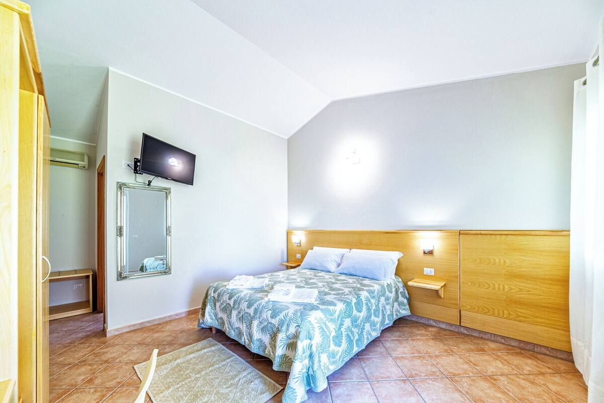 A bedroom with a bed, a TV mounted on the wall, a wooden headboard, and a nightstand with a lamp.
