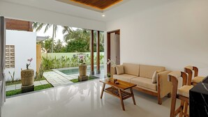 Ein modernes Wohnzimmer mit einem beigefarbenen Sofa, einem Holztisch und Sesseln. Ein großes Fenster bietet Blick auf einen Garten mit Palmen und einem Swimmingpool.