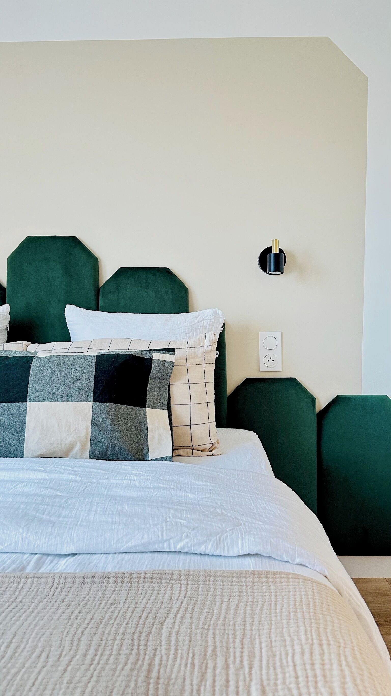 A neatly made bed with a white duvet and a checkered blanket, green upholstered headboards, and a wall-mounted light fixture.