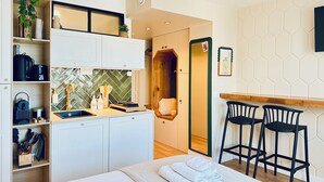 A modern hotel room with a kitchenette, a bed with neatly folded towels, a wooden chair, and a mirror. - L'Émeraude - Comfortable studio + private parking (Metz)