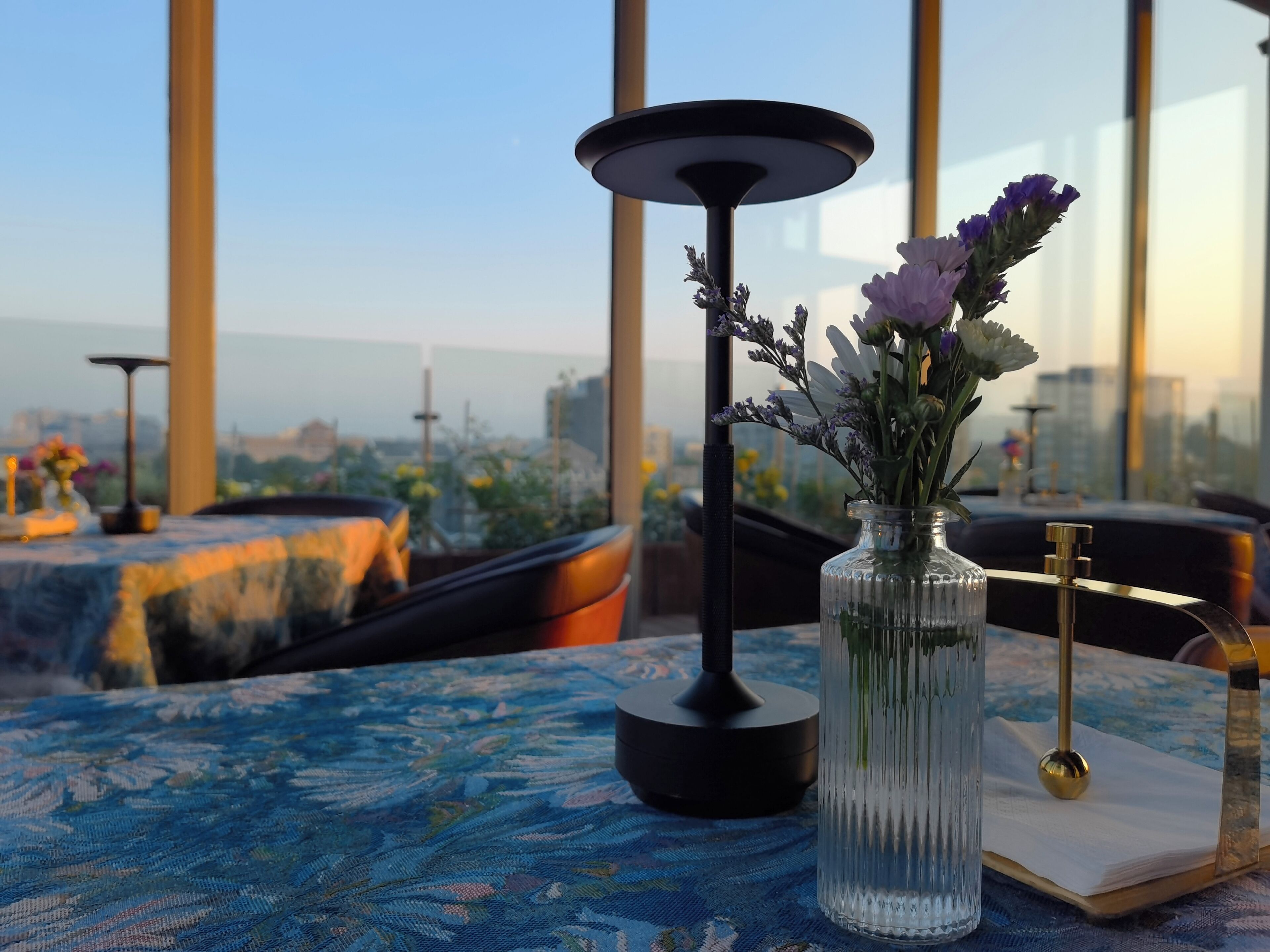 Une table ornée d’un vase de fleurs, d’une lampe et d’un livre, le tout posé sur une table recouverte d’une nappe à motifs bleus.