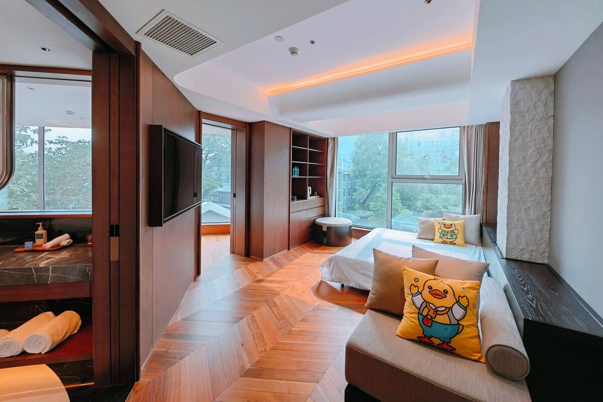A modern hotel room with a large bed, a sofa, a TV, and a view of the outdoors.