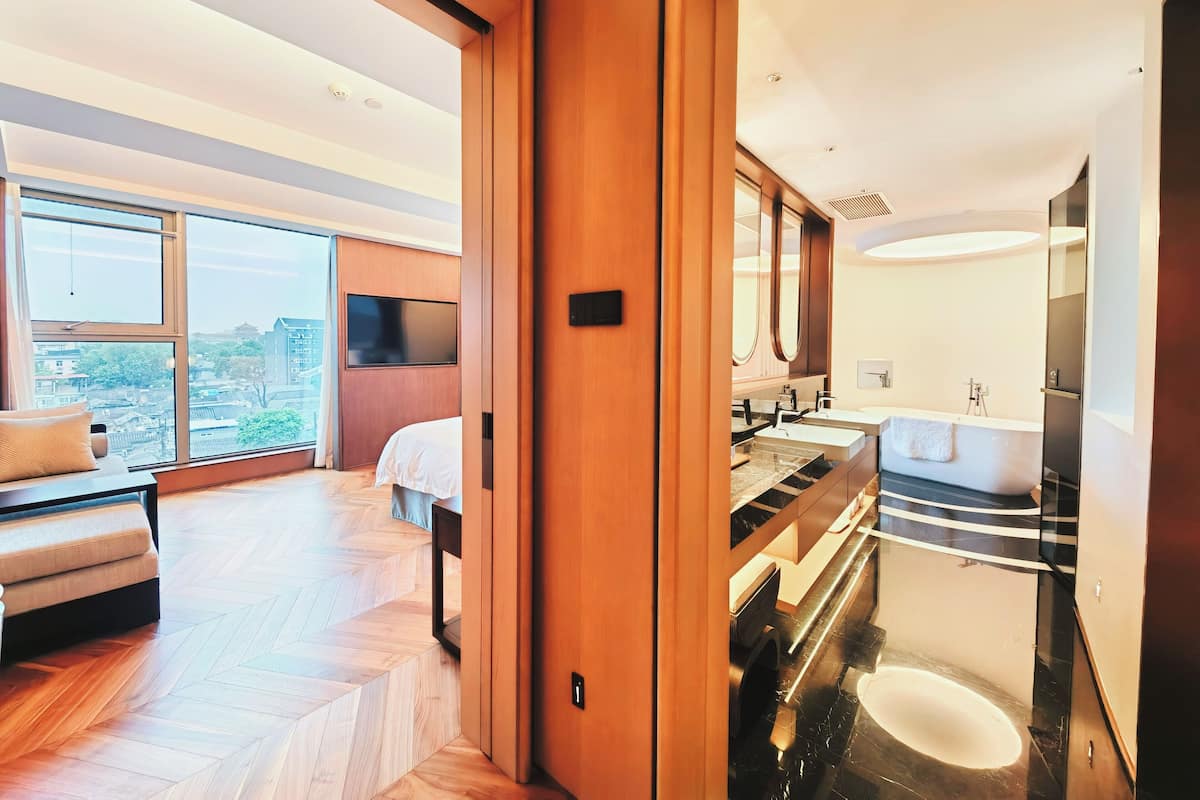 A modern hotel room with a large bed, a TV, a sofa, and a view of the city.