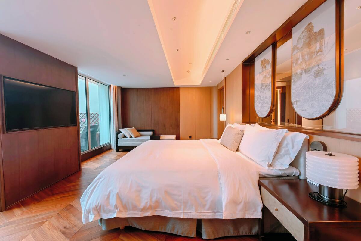 A modern hotel room with a large bed, a bedside table with a lamp, a TV mounted on the wall, and a large window offering a view of the outdoors.