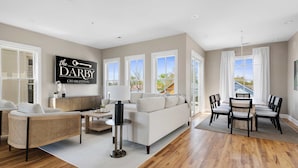 A spacious living room with a sofa, chairs, a coffee table, and a dining table with chairs. There is a large window with curtains, a wall-mounted sign, and a view of the outside.