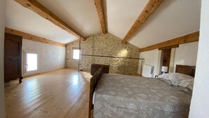 Une chambre avec un plafond en pente, des poutres en bois, un mur en pierre, un lit recouvert d’une couvre-lit à motifs floraux, une fenêtre et un radiateur.