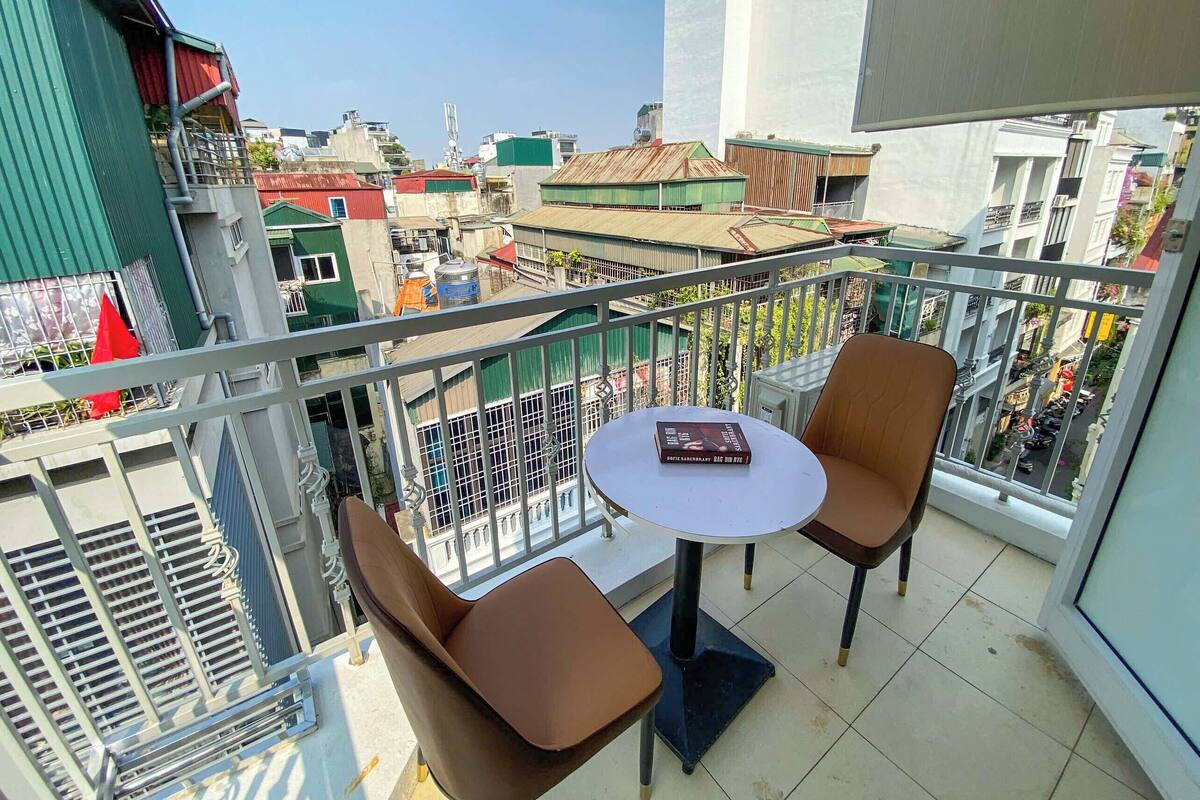 A balcony with two brown chairs and a small round table, overlooking a cityscape with various buildings and a clear sky.
