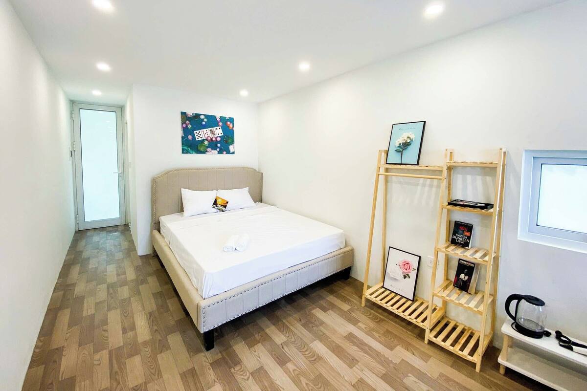 A bedroom with a bed, a wooden floor, a door, a wall-mounted shelf with books, and a framed picture.