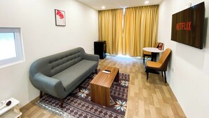 Una sala de estar moderna con un sofá gris, una mesa de centro de madera, un tapete estampado, una pequeña mesa redonda con una silla y un televisor montado en la pared.