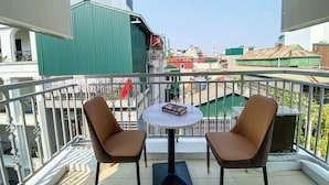 Un balcón con dos sillas y una mesa, con vista a un paisaje urbano que incluye edificios y una estructura con techo verde.