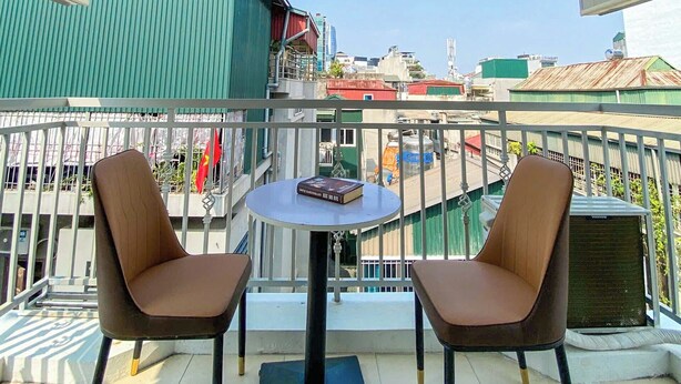 Un balcón con dos sillas y una mesita, con vistas a un paisaje urbano que incluye edificios y una bandera.