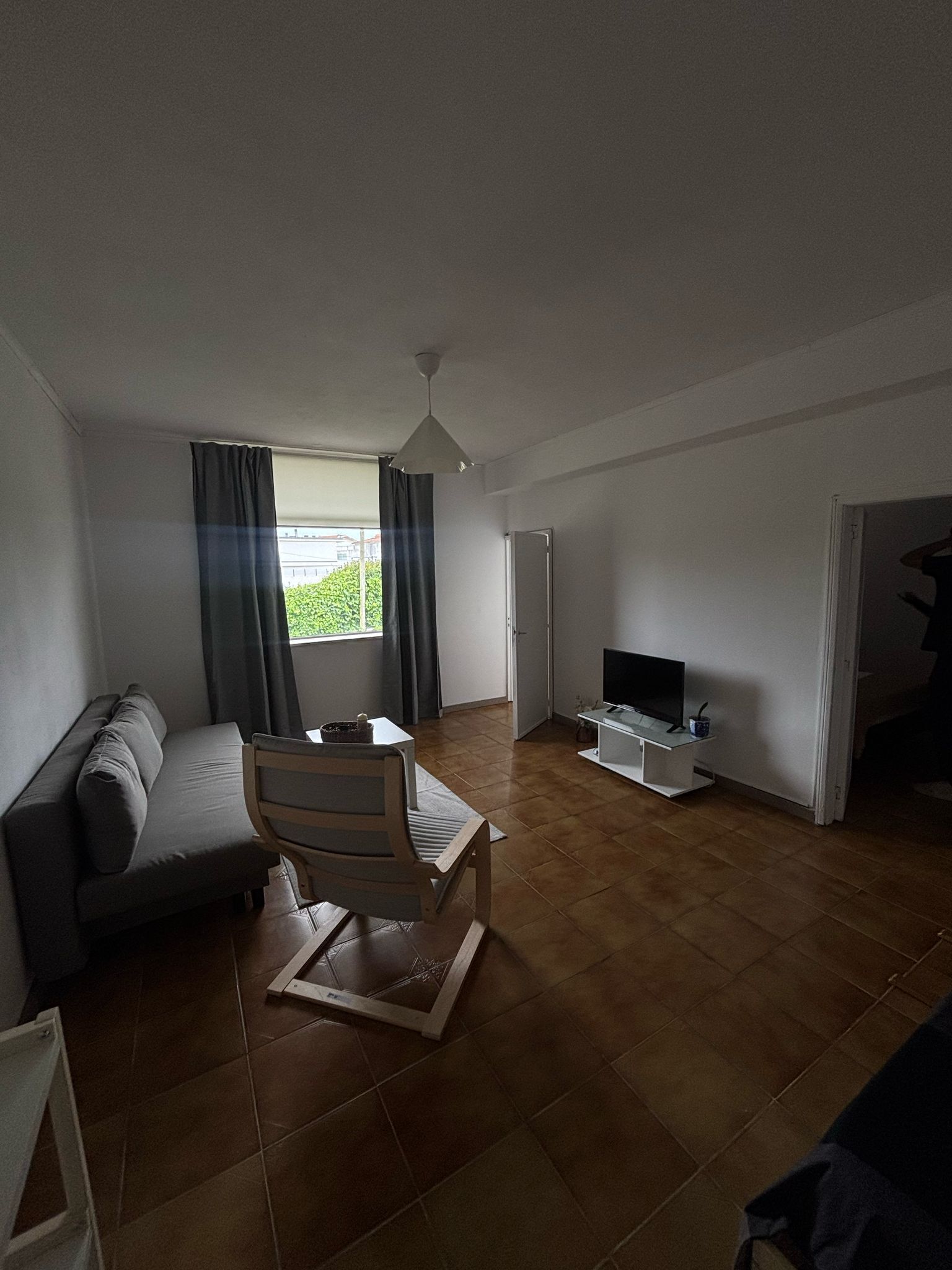Uma sala de estar moderna com sofá, uma cadeira de madeira, televisão e janela com cortinas.