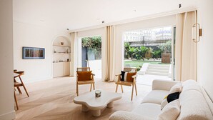 Een ruime woonkamer met een visgraatvloer, een witte bank, twee houten stoelen, een salontafel en uitzicht op een tuin door grote ramen.