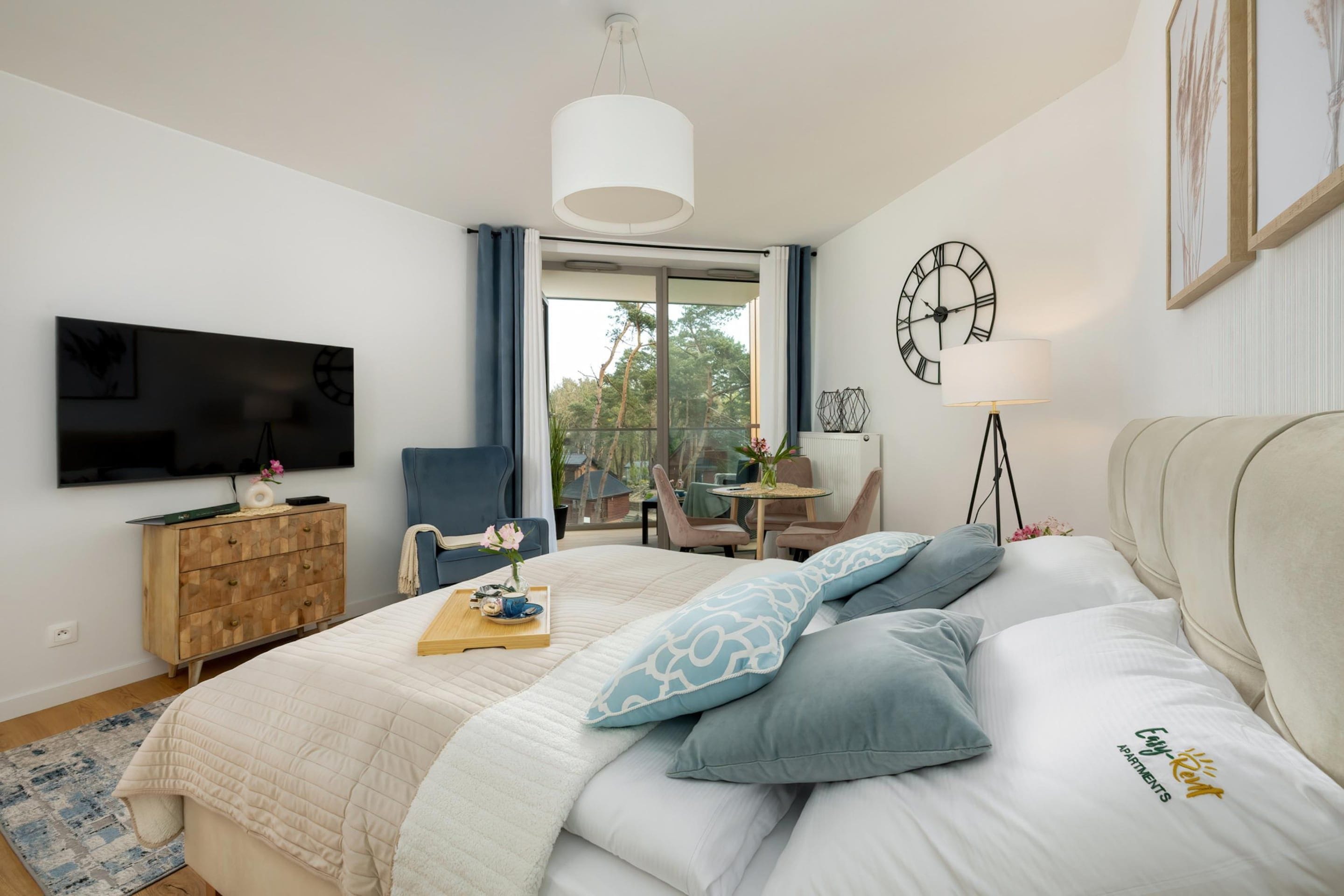 A bedroom with a bed, a TV, a desk with a chair, a window, and a clock on the wall.