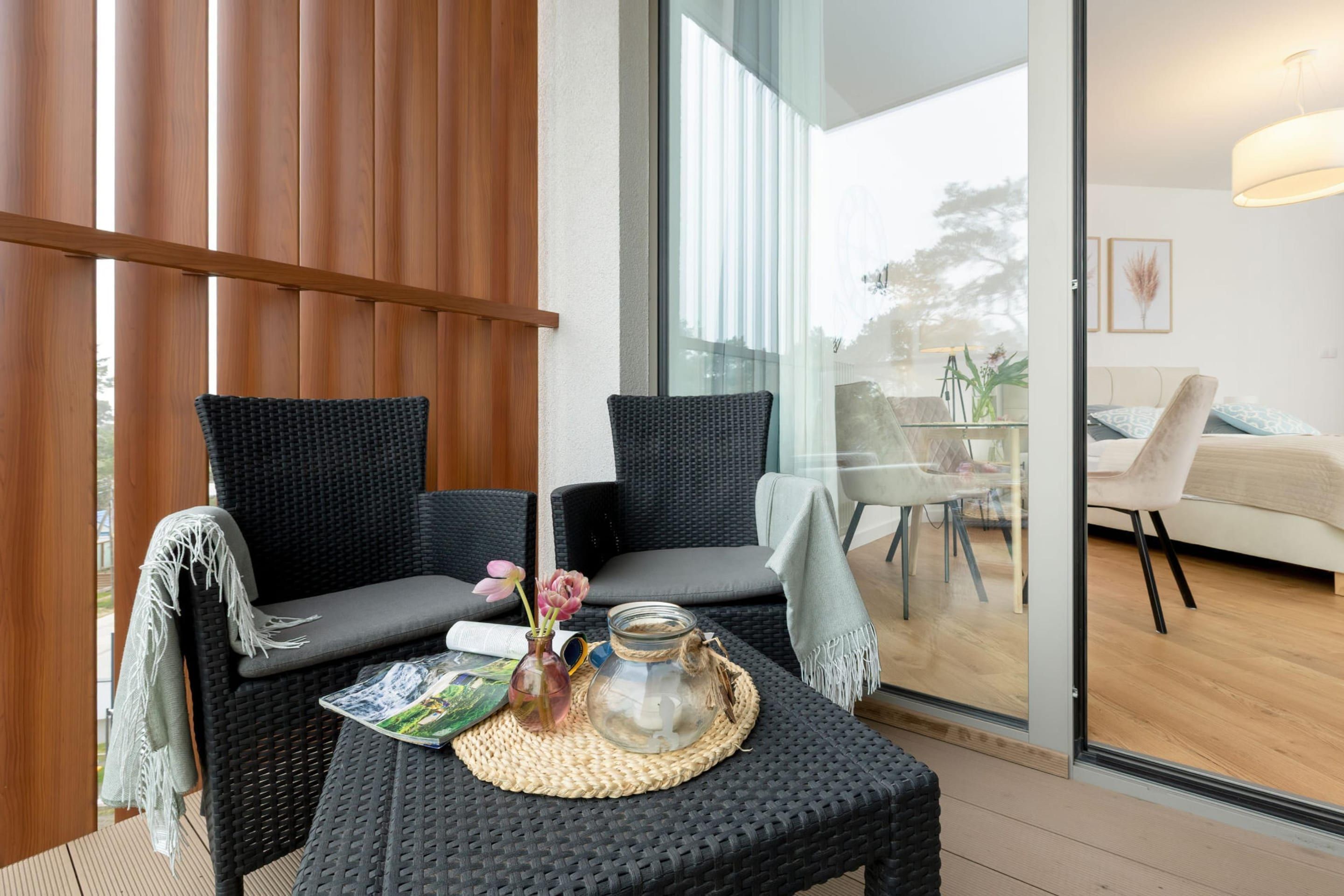Una habitación moderna con dos sillones de mimbre negro, una mesita con un mantel tejido, un florero con flores y un tarro de vidrio.