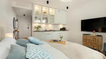 Un dormitorio moderno con cama, almohadas, una mesita de noche, televisor, un armario de madera y una zona de cocina con muebles y encimera.