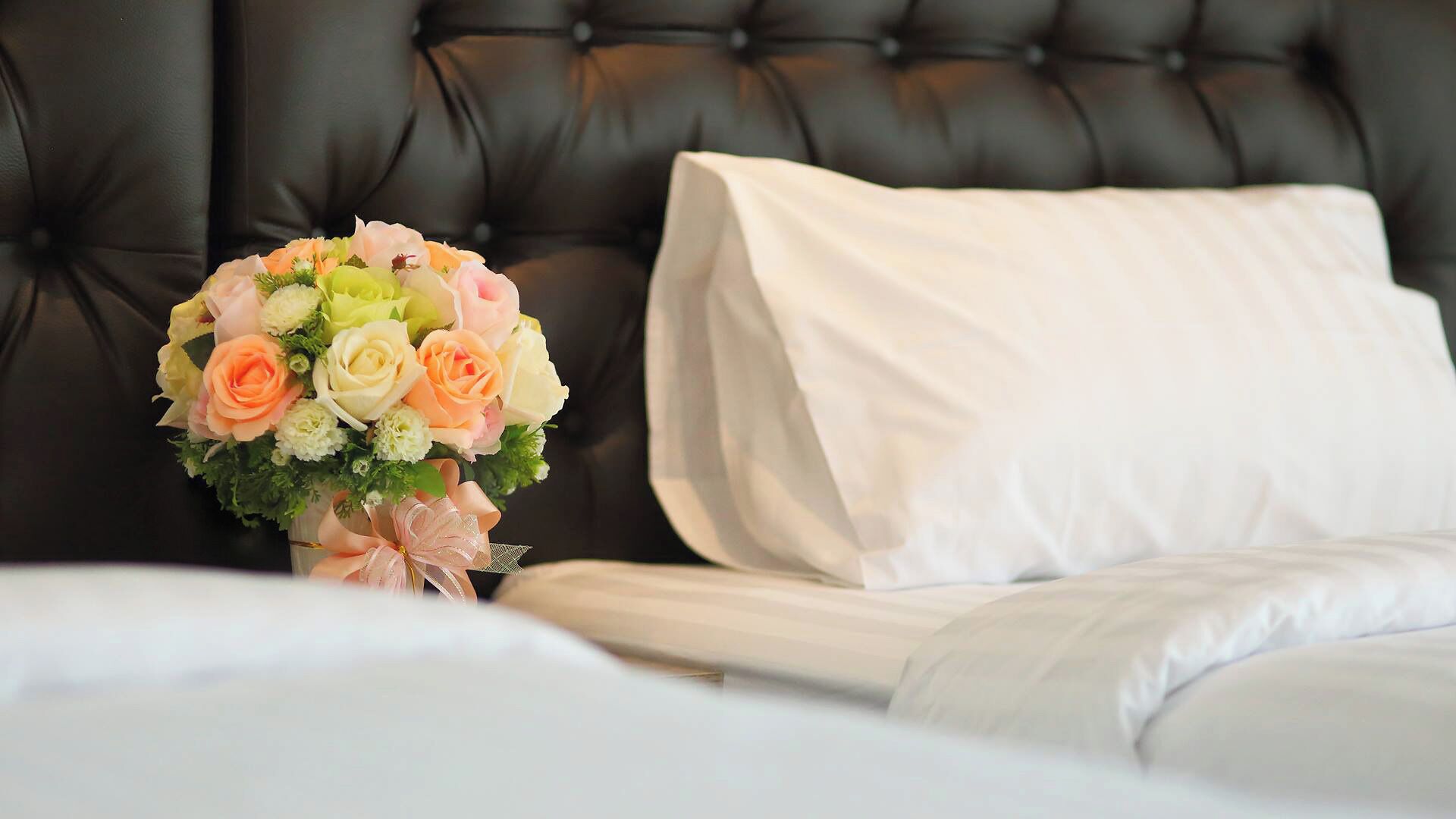 A bed with white linens, a bouquet of flowers, and a black upholstered headboard.