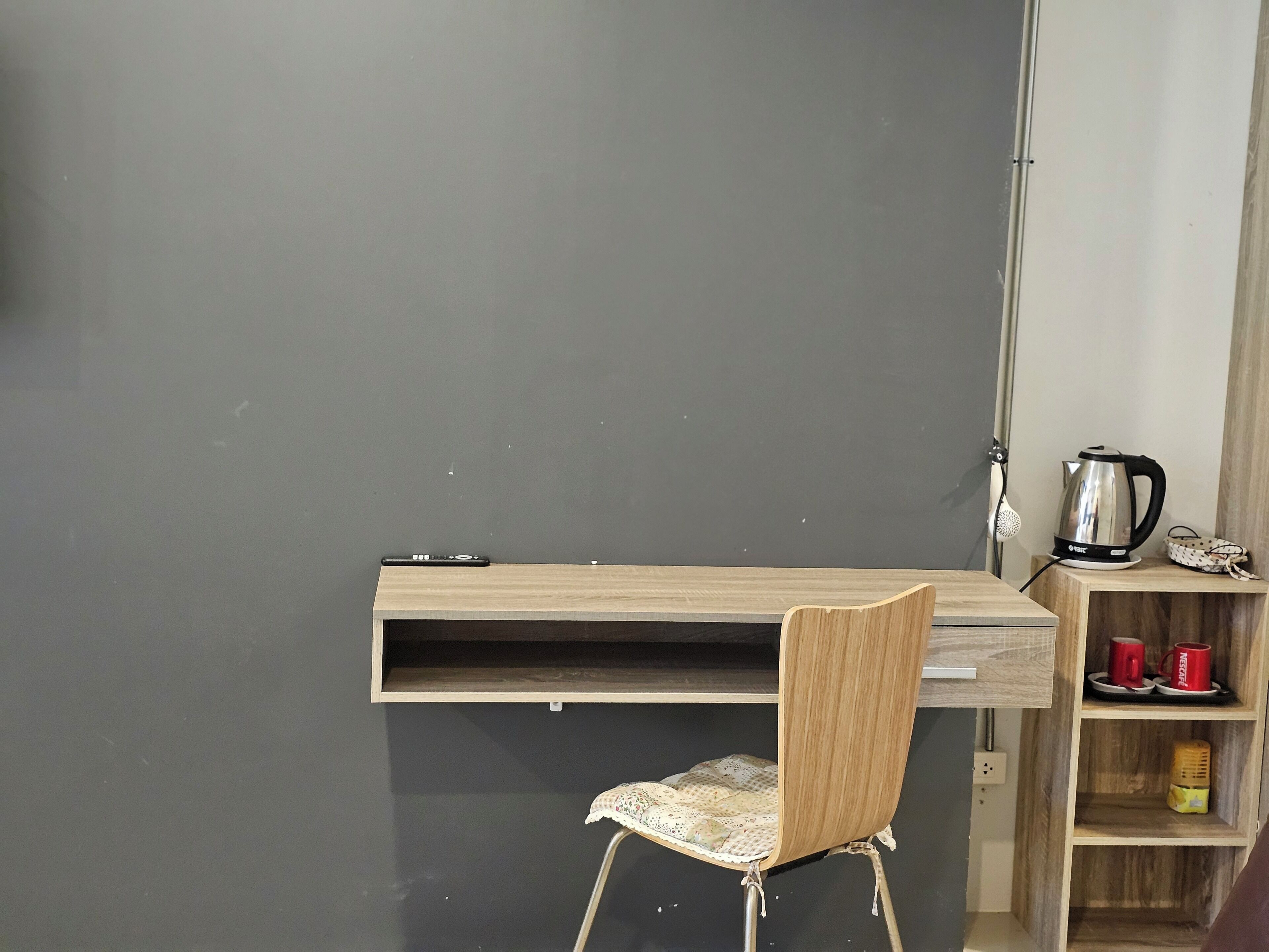 A room with a desk, chair, and a shelf with a kettle and mugs.