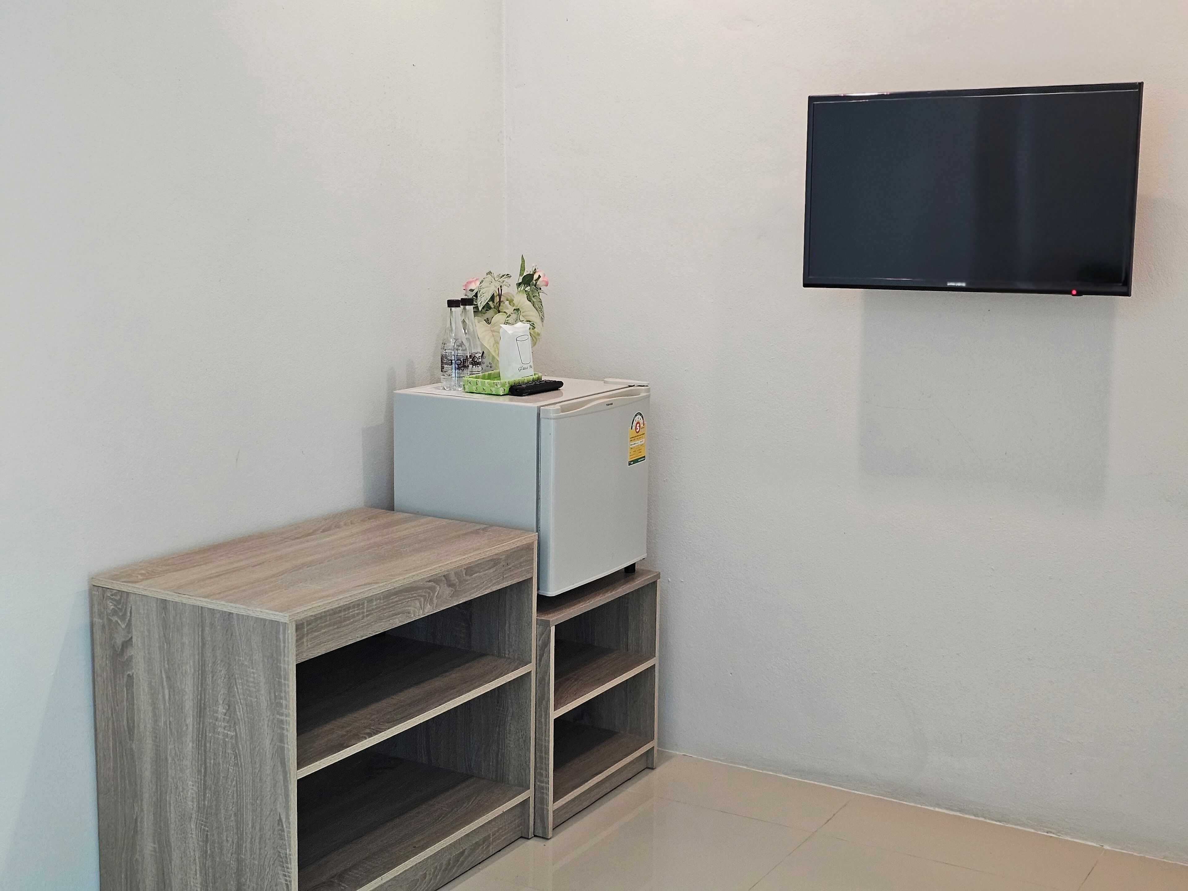 A small room with a white wall, a mounted TV, a white mini-fridge, and a wooden shelf unit.
