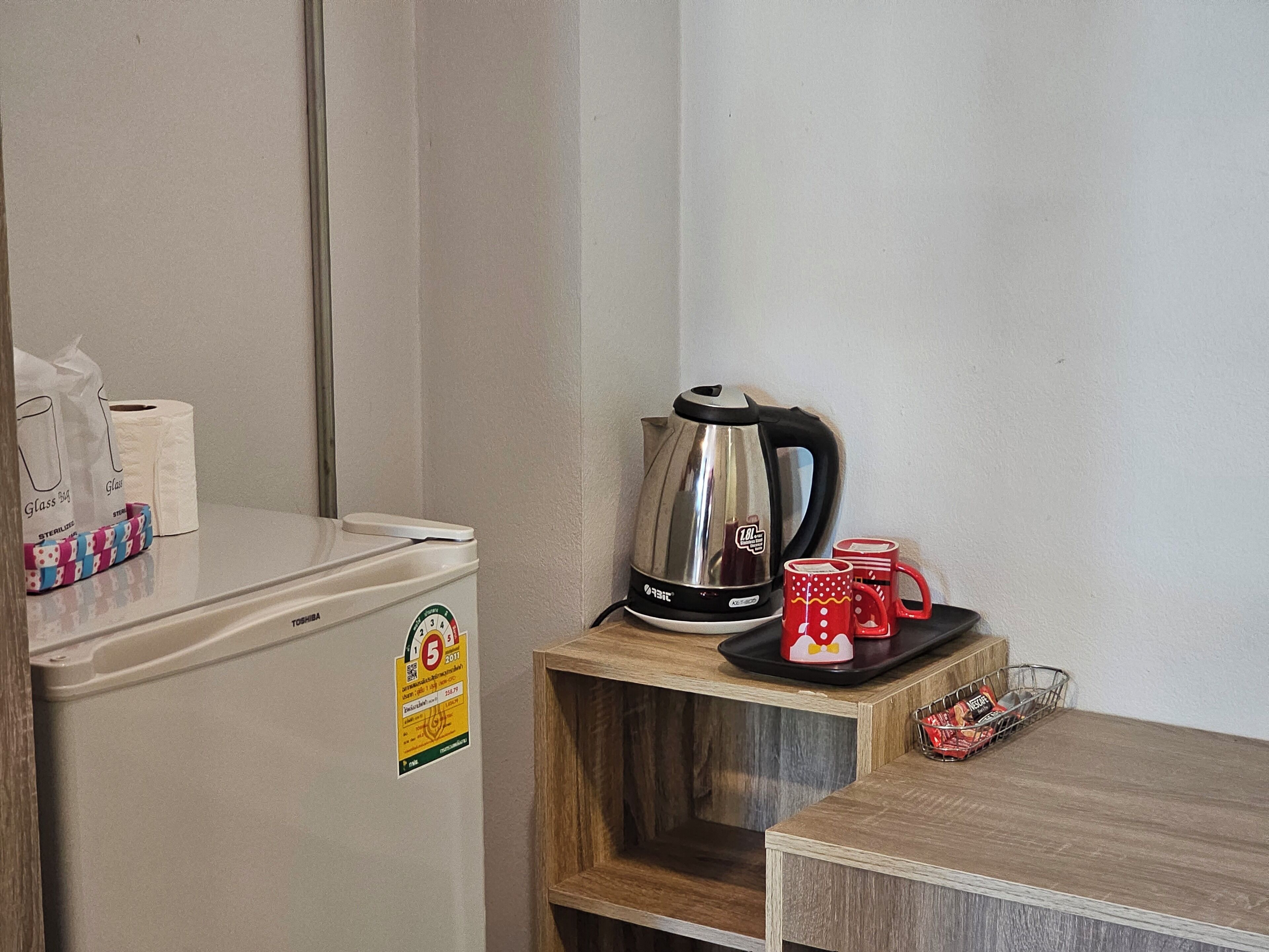 A kitchenette with a refrigerator, a kettle, and two mugs on a wooden shelf.