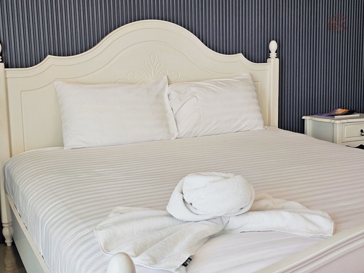 A neatly made bed with white linens and pillows, a white headboard, and a small bedside table with a book and a remote control.