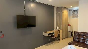 A modern hotel room with a flat-screen TV mounted on the wall, a wooden desk with a chair, a wardrobe, a brown leather sofa, and a neatly made bed with a rolled towel.