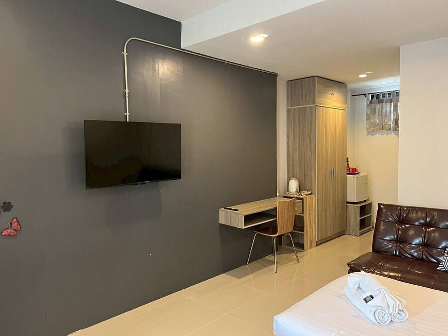 A modern hotel room with a flat-screen TV mounted on the wall, a wooden desk with a chair, a wardrobe, a brown leather sofa, and a neatly made bed with a rolled towel.