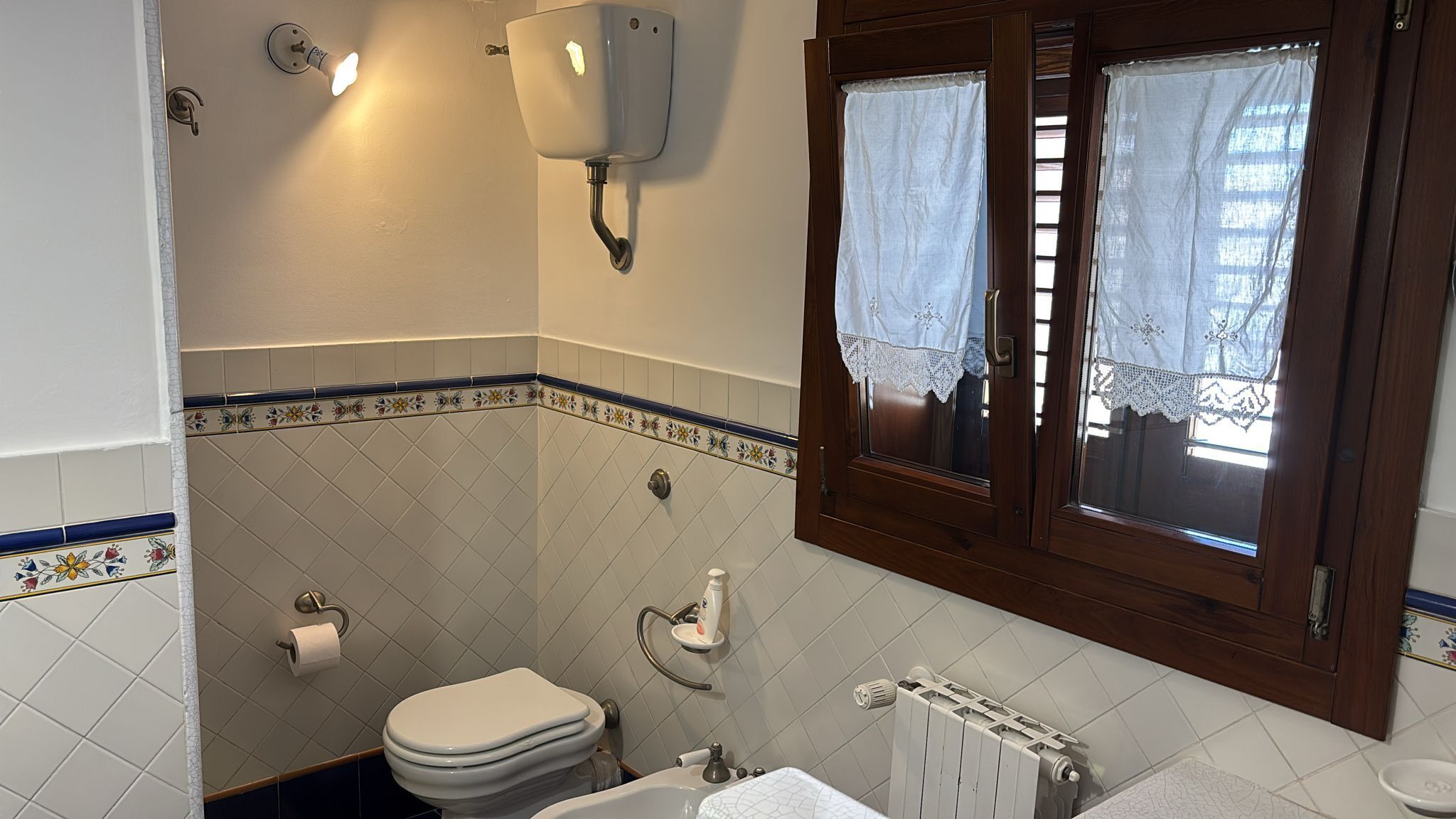 Une salle de bain avec des toilettes, un lavabo et une fenêtre ornée de rideaux en dentelle.