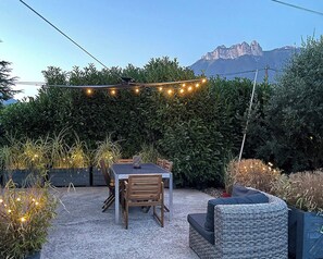 An outdoor patio with a table and chairs, surrounded by greenery and string lights.
