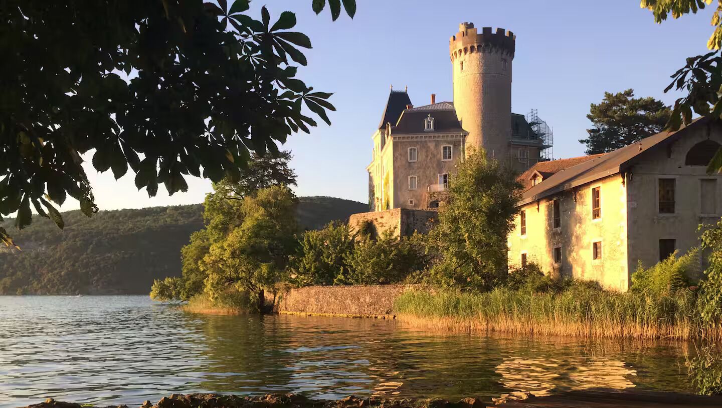 A castle with a tower by a lake, surrounded by trees and a stone wall.