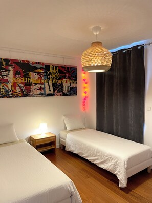A bedroom with two beds, a wooden nightstand, a wall-mounted lamp, a hanging light fixture, and a colorful abstract painting.
