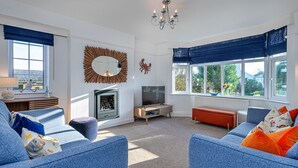 A living room with a fireplace, a television, and a large window with blue curtains. - House in Abersoch Near Sandy Beach (Pwllheli)