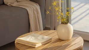 Ett modernt vardagsrum med en grå soffa, ett träbord med en bok på och en vas med gula blommor.
