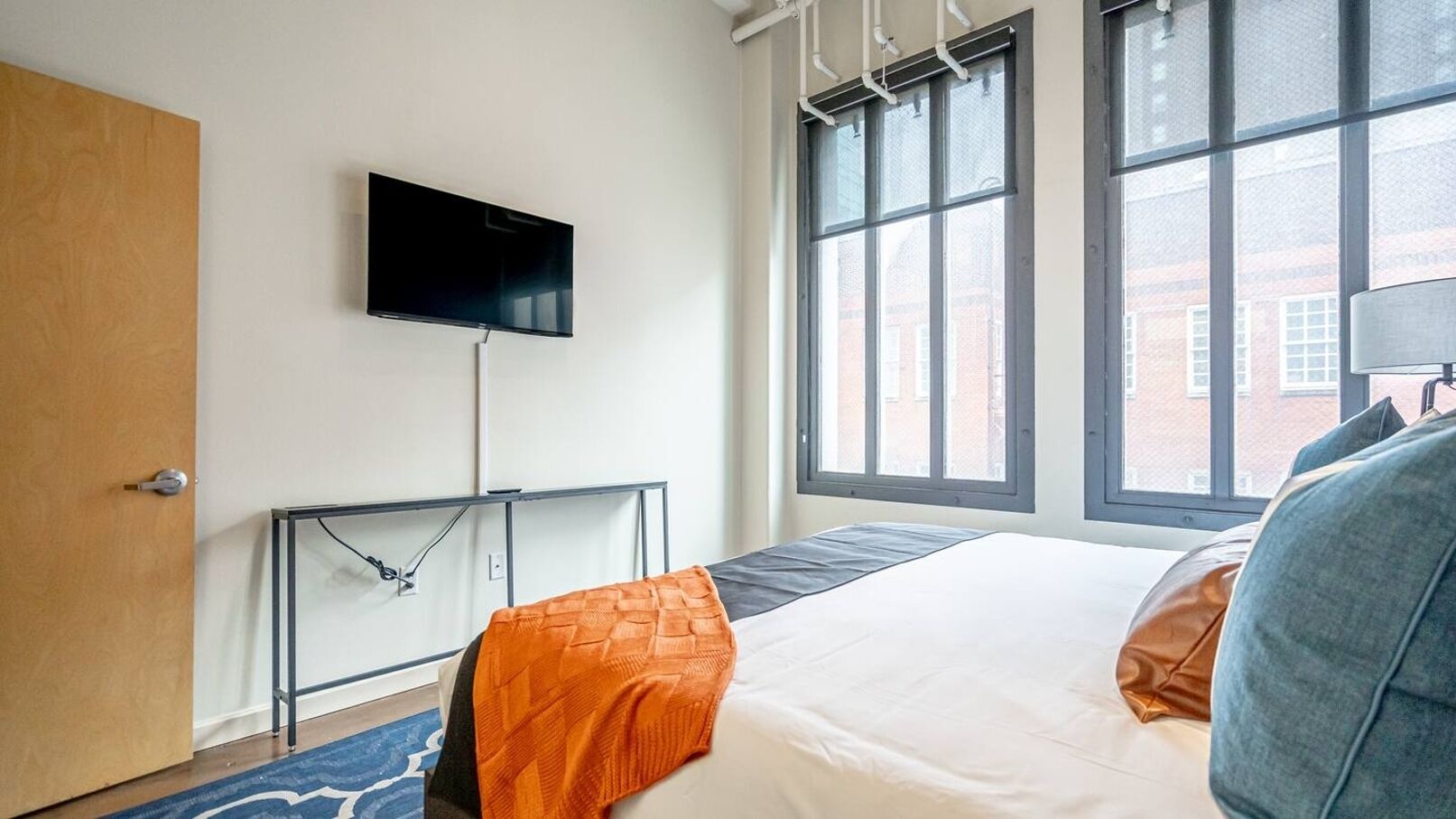 A hotel room with a bed, a TV mounted on the wall, a window with a view of buildings, and a door.