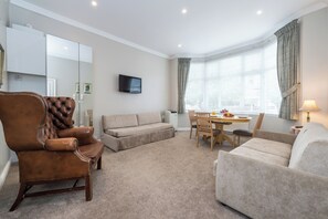 A living room with a brown leather armchair, beige sofa, and a small dining table with chairs. A flat-screen TV is mounted on the wall, and there is a window with curtains. - Beautiful Apartment ac up to Six,12 min by Tube From Oxford Street,ac (London)