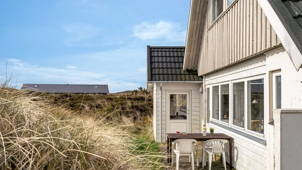 Una casa costera con terraza de madera, una pequeña mesa con sillas y una puerta que da al interior.