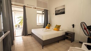 Hotelkamer met een bed, nachtkastje, ventilator en uitzicht op een zwembad en palmbomen.