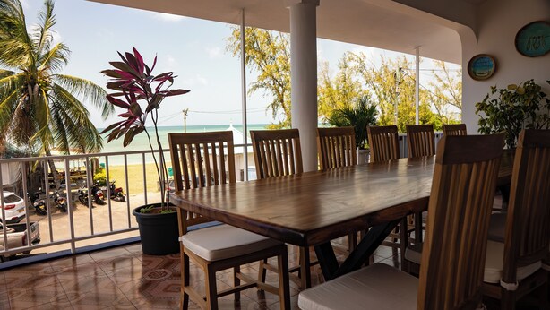 Een houten eettafel met stoelen, een plant in pot en uitzicht op het strand en palmbomen.