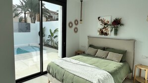 A bedroom with a bed, a small table, a wall-mounted light fixture, and a view of a swimming pool through a sliding glass door.