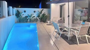 A swimming pool with a glass table and chairs, surrounded by plants and a patio area.