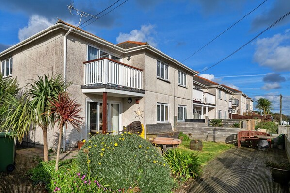 A two-story residential building with a balcony, a small garden with various plants, and a wooden deck. - Apartment in Newquay Near Stunning Beaches (Newquay)