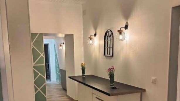 Un couloir qui mène à un espace cuisine, avec des meubles de couleur claire, un plan de travail foncé et des luminaires fixés au mur.