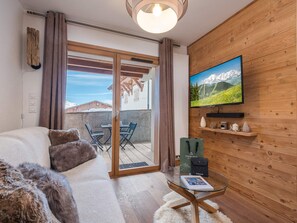 A cozy living room with a sofa, a glass coffee table, a TV mounted on a wooden wall, and a sliding glass door leading to an outdoor patio. - Cozy apartment near slopes, 4 pax, Les Saisies (Les Saisies)