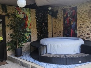 A circular hot tub with a white cover, surrounded by a black wicker seating area, set against a stone wall with a tropical mural and a potted plant. - Cottage for 8 with Fireplace and SPA in Biron (Biron)