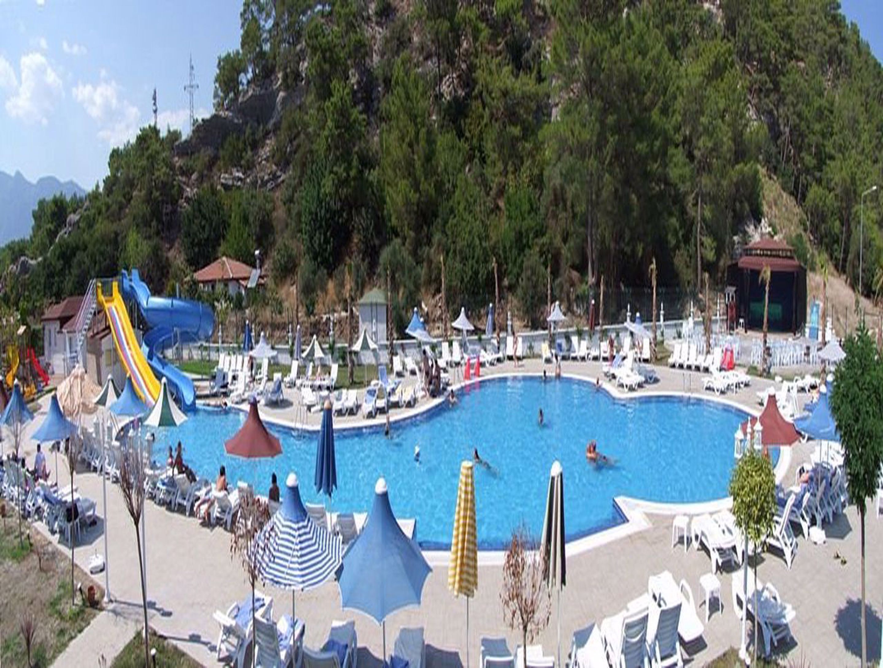 Zona de piscina con sillas de descanso y sombrillas, rodeada de árboles y una colina al fondo.