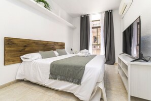 Moderni hotellihuone, jossa on puinen pääty, valkoisilla lakanoiden ja vihreän huovan peittämä sänky, taulutelevisio ja parveke, jossa on verhot.