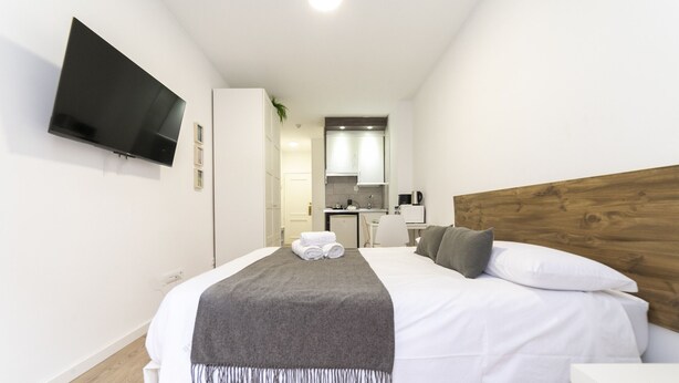 Habitación de hotel con cama, televisión montada en la pared, kitchenette y cabecera de madera.